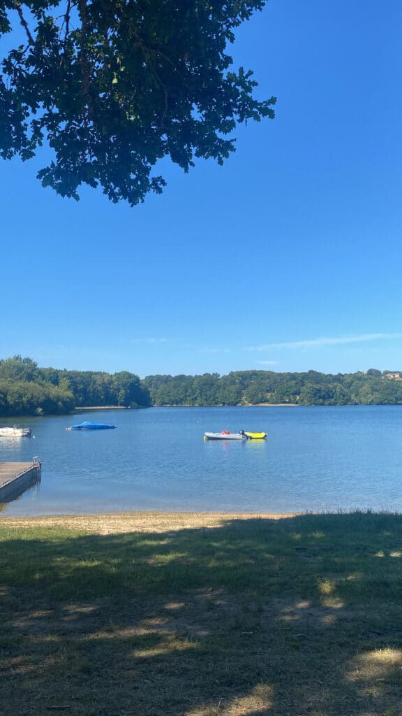 Une scène paisible au bord d'un lac, sous un ciel bleu limpide. Deux personnes à bord de pédalos flottent sur l'eau calme, entourées d'une végétation luxuriante. Une atmosphère de détente et de sérénité.