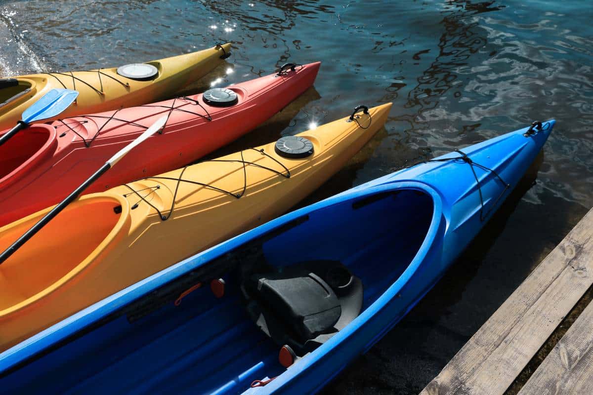 Trois kayaks aux couleurs vives — bleu, jaune et rouge — sont alignés côte à côte sur un ponton en bois, flottant paisiblement sur une eau légèrement agitée.