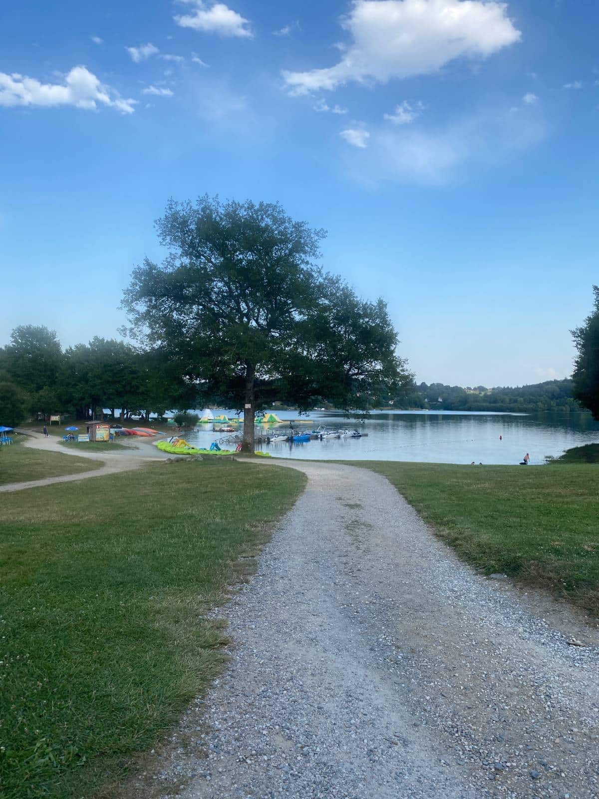 Une scène paisible au bord d'un lac, avec un chemin de gravier menant à un arbre près de l'eau. Des kayaks et des pédalos colorés sont visibles sous un ciel bleu éclatant.
