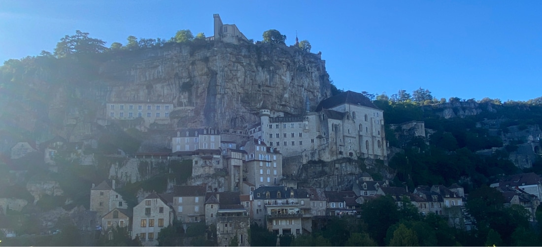 Une vue pittoresque d'un village à flanc de colline, bâti à même une falaise rocheuse et composé de bâtiments médiévaux en pierre sous un ciel bleu limpide. La scène est empreinte de sérénité et d'histoire.