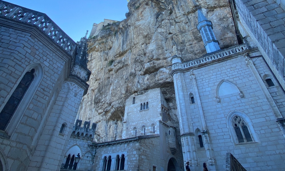 Une architecture spectaculaire de style gothique se dressant face à une falaise abrupte et imposante, sous un ciel bleu limpide. La scène évoque un sentiment d'émerveillement et de grandeur historique.