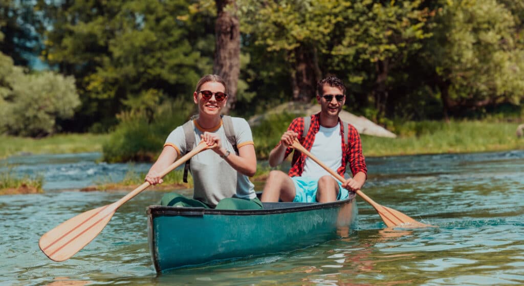 Un couple souriant portant des lunettes de soleil fait du canoë sur une rivière calme et limpide, entouré d'une végétation luxuriante par une journée ensoleillée, dégageant une atmosphère joyeuse et empreinte d'aventure.