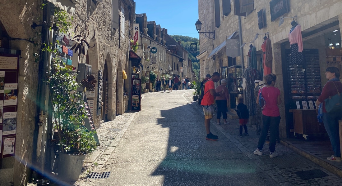 Une rue étroite pavée, bordée de bâtiments en pierre et de petites boutiques. Quelques personnes flânent tandis que la lumière du soleil projette des ombres, créant une atmosphère chaleureuse et accueillante.