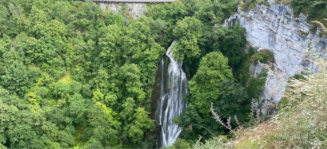 Une cascade paisible dévale une falaise verdoyante, entourée d'un feuillage dense et de formations rocheuses. La scène évoque la tranquillité et la beauté naturelle.