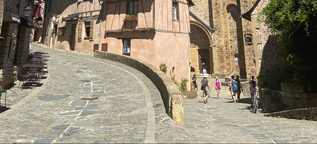 Une rue pavée serpente doucement entre des bâtiments rustiques en pierre et en bois agrémentés de jardinières fleuries. Des touristes flânent, profitant du cadre historique ensoleillé.