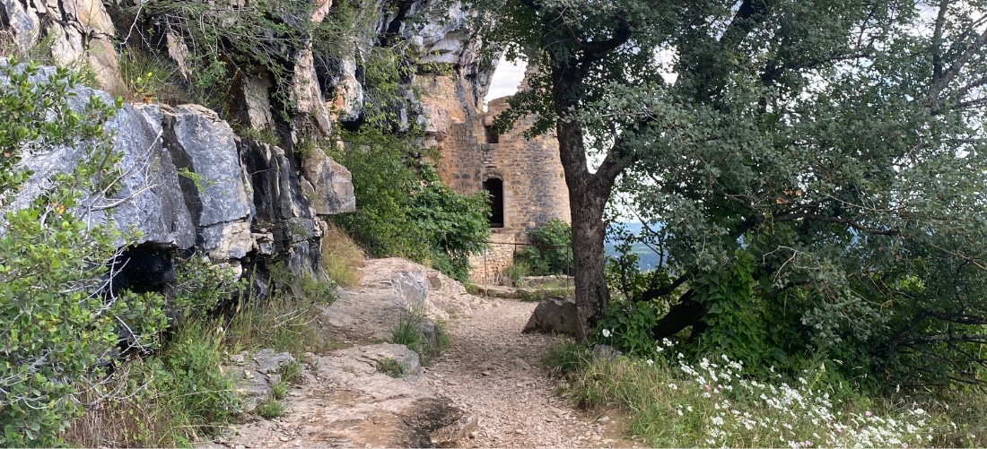 Un sentier accidenté mène à une ancienne construction en pierre nichée dans des falaises rocheuses, entourée d'une végétation luxuriante et d'arbres, évoquant un sentiment de mystère et de solitude.