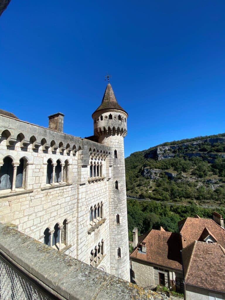 Une tour de château en pierre, dotée de fenêtres en plein cintre et d'un toit conique, se dresse sous un ciel bleu limpide. À proximité, des bâtiments traditionnels et des collines verdoyantes complètent le tableau.