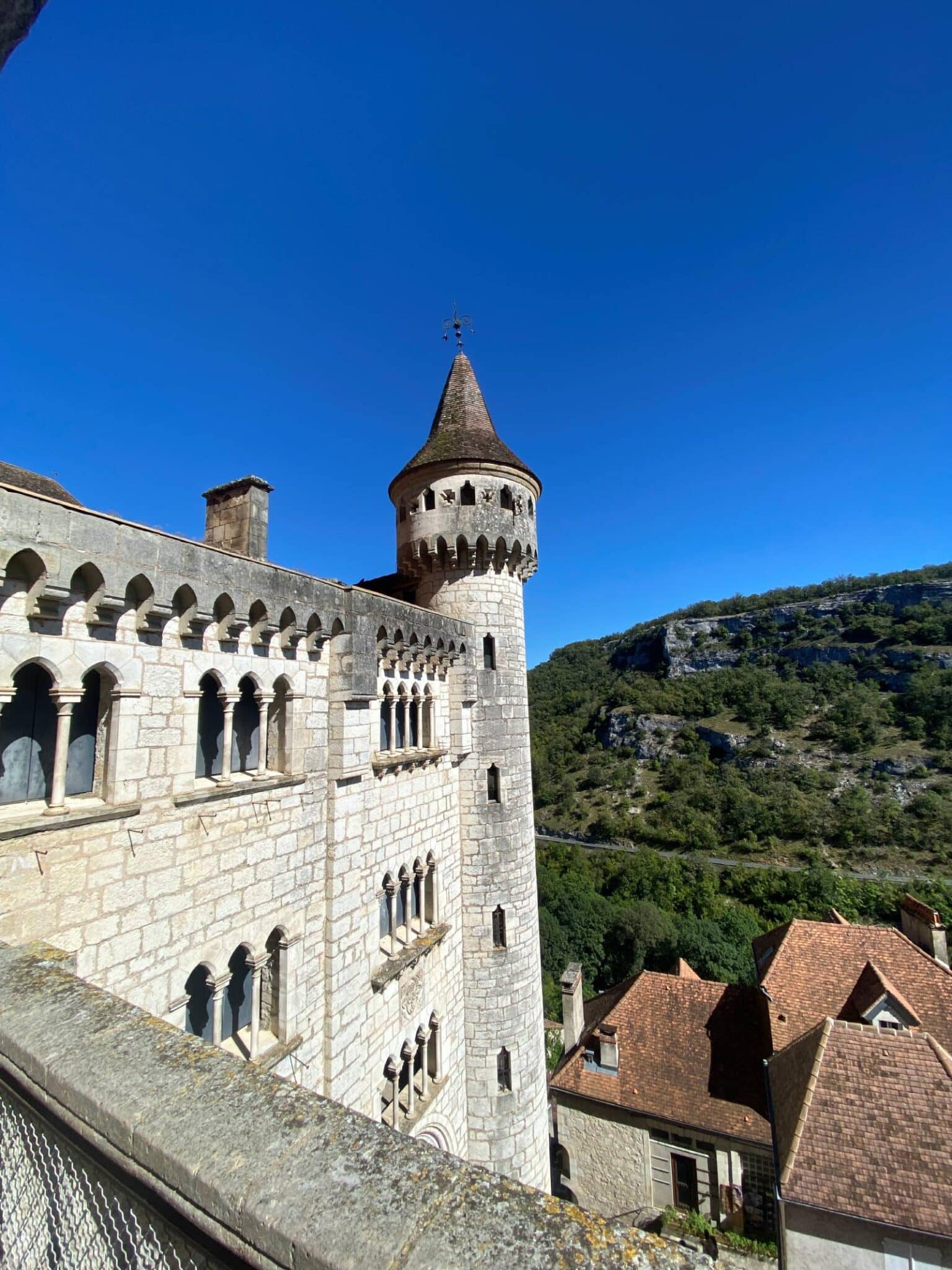 Une tour de château en pierre, dotée de fenêtres en plein cintre et d'un toit conique, se dresse sous un ciel bleu limpide. À proximité, des bâtiments traditionnels et des collines verdoyantes complètent le tableau.
