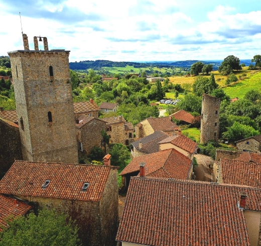Vue aérienne d'un village pittoresque aux tours de pierre et aux toits de tuiles rouges, entouré d'une végétation luxuriante et de collines vallonnées sous un ciel partiellement nuageux.