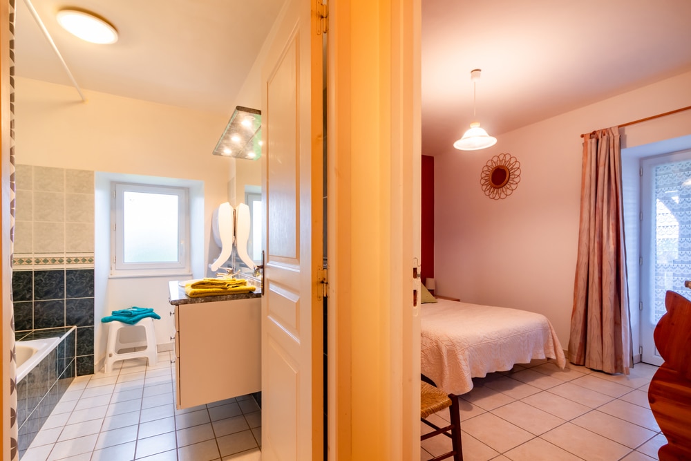 Vue chaleureusement éclairée d'une salle de bains et d'une chambre séparées par une porte entrouverte. La salle de bains comprend une baignoire et un lavabo ; la chambre est équipée d'un lit, d'une chaise et d'une fenêtre. Ambiance chaleureuse.