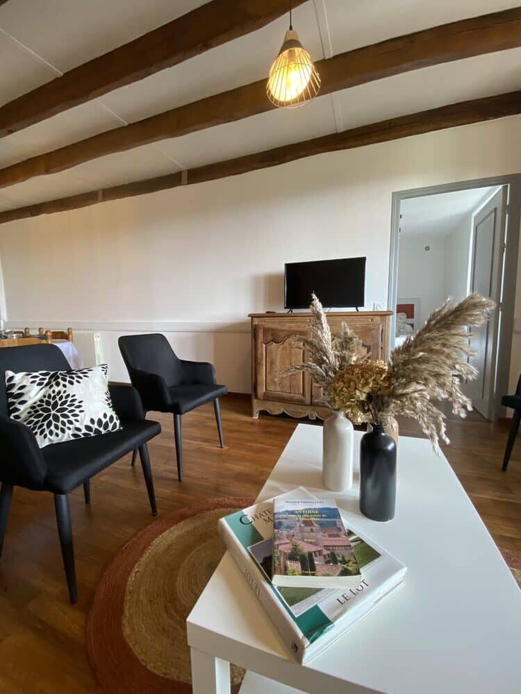 Un salon chaleureux doté de plafonds aux poutres apparentes et d'un éclairage doux. Des chaises noires agrémentées de coussins à motifs sont disposées près d'un téléviseur posé sur un meuble en bois. Une table blanche accueille des fleurs séchées et des livres, créant une atmosphère accueillante.