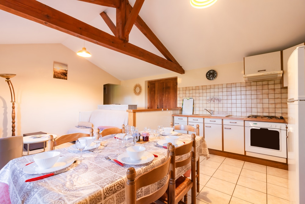 Un espace cuisine et salle à manger chaleureux, doté d'un plafond aux poutres apparentes. La table est dressée avec de la vaisselle et des couverts. Un éclairage chaleureux crée une ambiance accueillante.