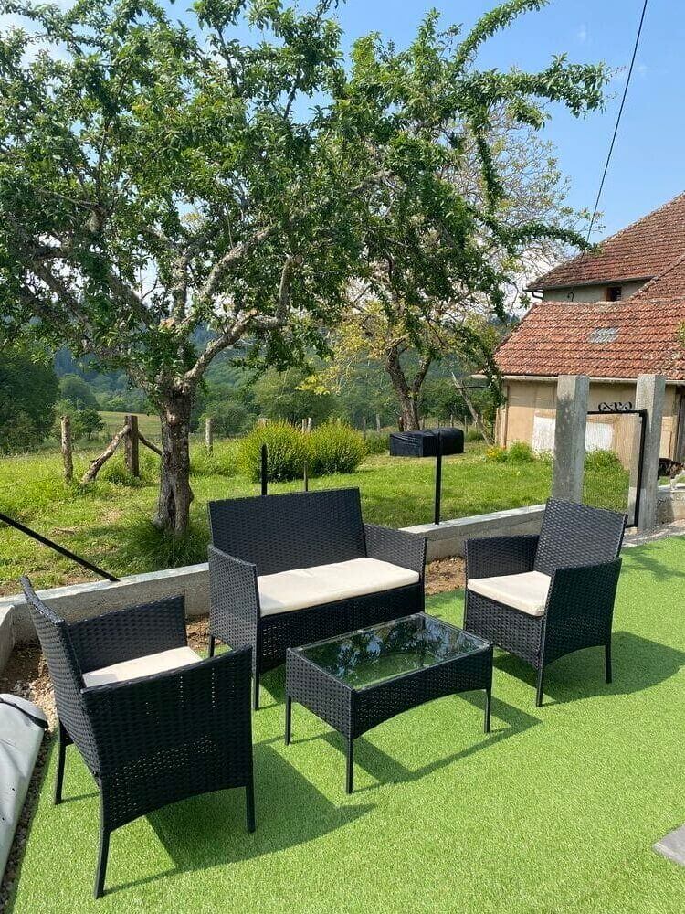 Espace de détente en plein air avec du mobilier en osier noir sur du gazon artificiel vert. Un arbre et une maison au toit rouge se trouvent en arrière-plan, sous un ciel dégagé.