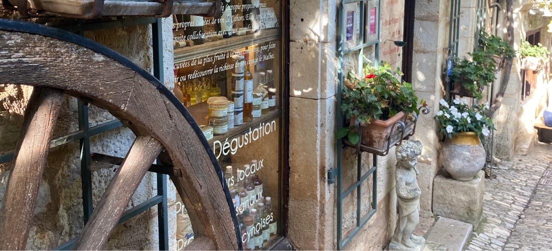 La façade rustique d'une boutique, avec une roue de charrette en bois, un mur en pierre et une vitrine exposant bouteilles et bocaux. Des pots de fleurs et une petite statue ajoutent du charme. Un chemin pavé baigné de soleil.