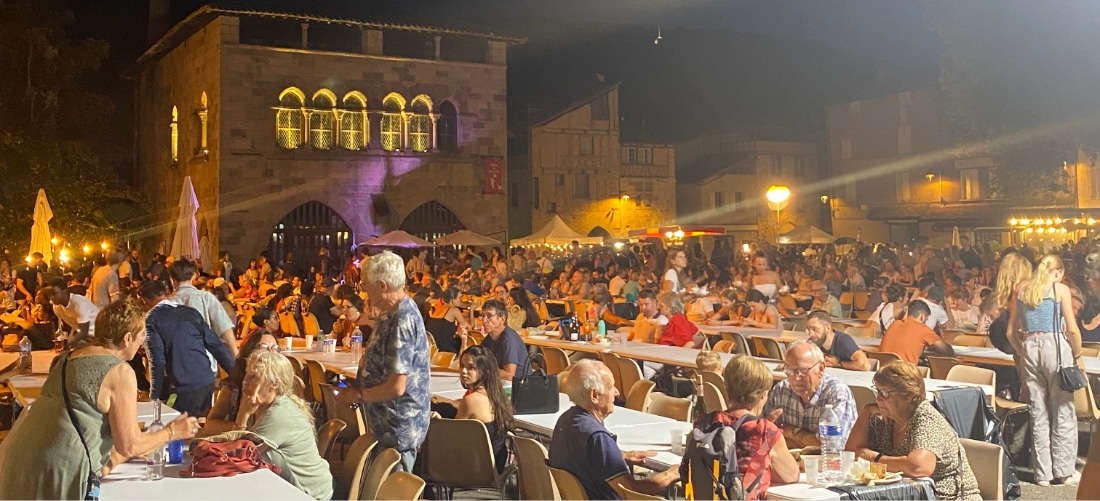 Un festival nocturne animé en plein air, avec une foule attablée et baignée dans une lumière chaleureuse. Un bâtiment historique en arrière-plan ajoute au charme des lieux.