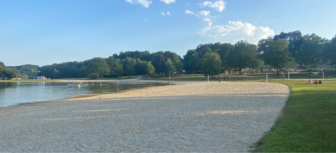 Une scène de plage paisible où le sable fin s'étend jusqu'aux eaux calmes d'un lac, sur la gauche. De l'herbe verte borde la droite, et des arbres sous un ciel bleu ajoutent à la sérénité du lieu.