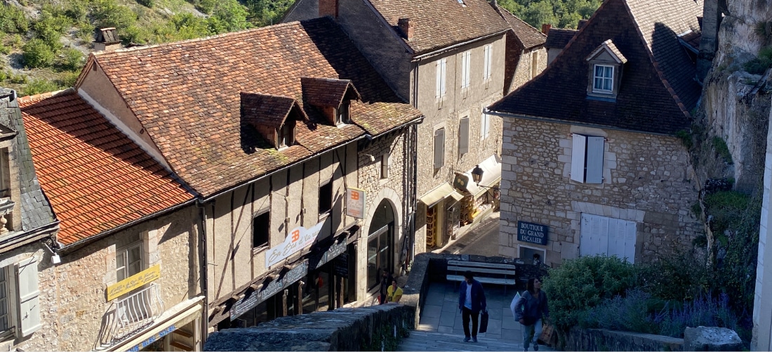 Village européen historique aux bâtiments en pierre et aux toits rouges rustiques. Une rue étroite et en pente présente des personnes descendant des escaliers, entourées de verdure.
