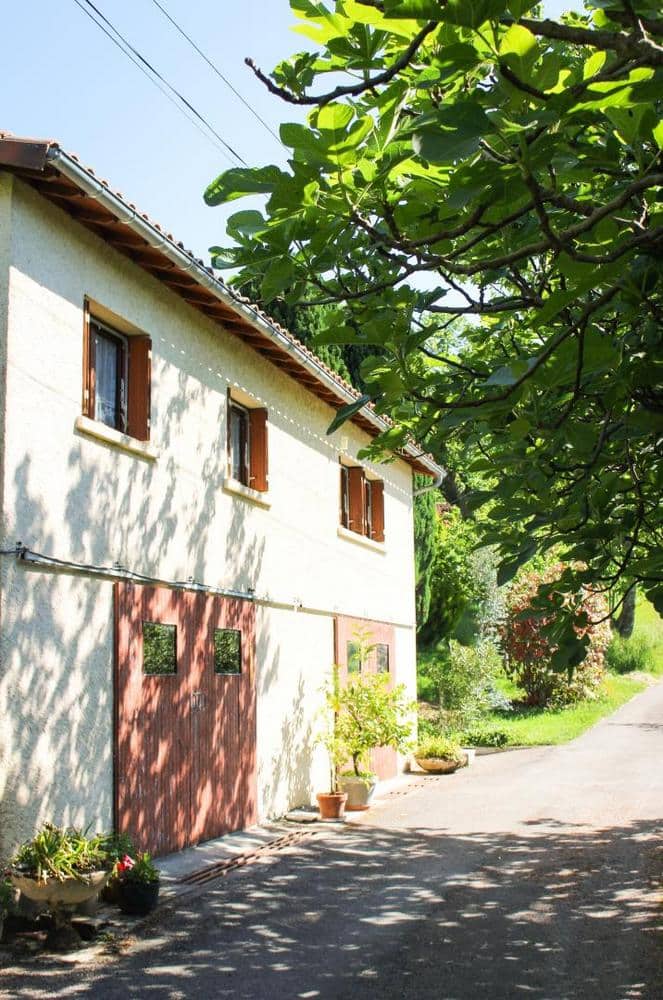Une maison rustique à deux étages, dotée de volets en bois et d'un toit en tuiles d'argile, est baignée de soleil. Un arbre projette des ombres mouchetées, créant une atmosphère sereine et accueillante.
