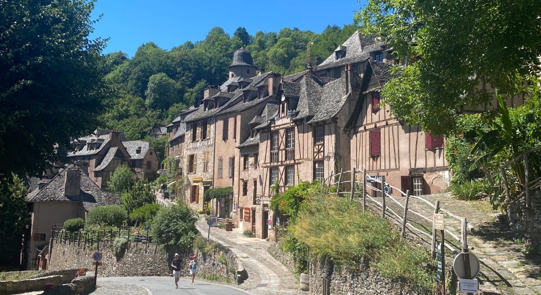 Une scène charmante de village médiéval avec des rues pavées et des maisons rustiques aux façades de pierre et de bois, le tout entouré d'une végétation luxuriante par une journée ensoleillée.