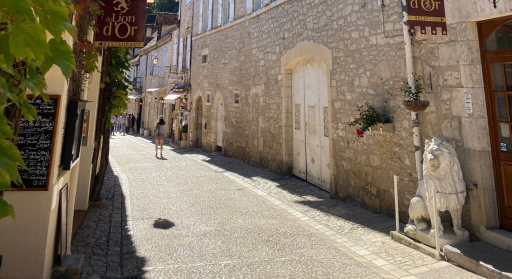 Rue pavée dans un village européen baigné de soleil, bordée de bâtiments en pierre. Une sculpture de lion et des fleurs ornent l'entrée d'un restaurant.