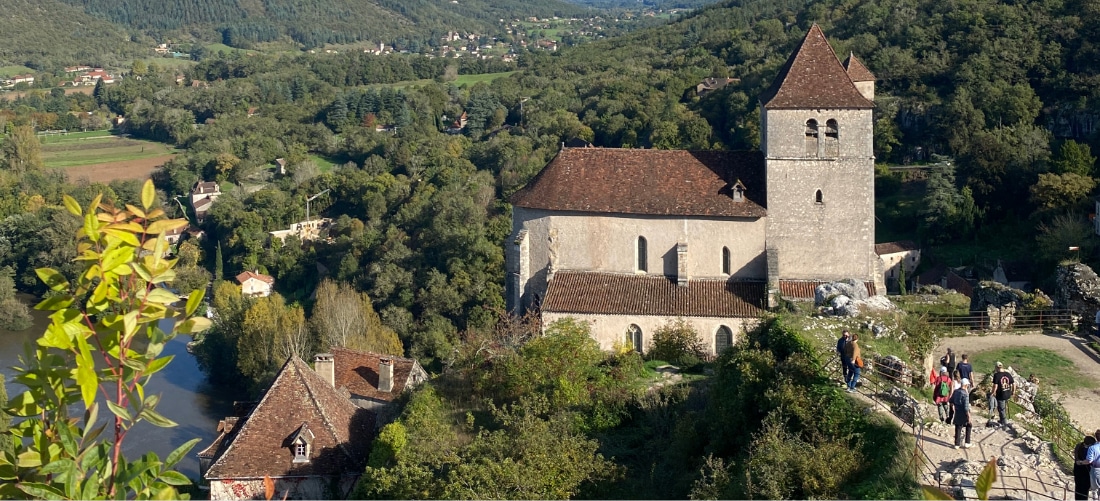 Vue aérienne d'une église médiévale en pierre, dotée d'un haut clocher et d'un toit en tuiles rouges, entourée d'une forêt verdoyante et de collines vallonnées sous un ciel bleu limpide. Des touristes se promènent sur un sentier voisin, conférant une atmosphère animée au lieu.