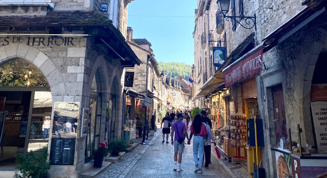 Une rue pittoresque aux pavés, dans un village européen, bordée de bâtiments en pierre, de boutiques et d'enseignes colorées. Des gens se promènent tranquillement sous un ciel bleu limpide.