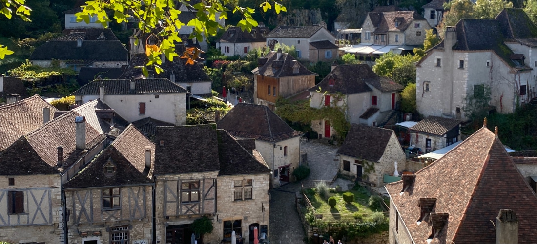Un village médiéval pittoresque aux maisons de pierre, aux toits pentus et au lierre. Un feuillage baigné de soleil encadre la scène, évoquant un charme serein et intemporel.